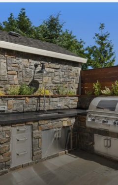 An outdoor kitchen features a stone wall, stainless steel grill, sink, and cabinets under a clear blue sky.