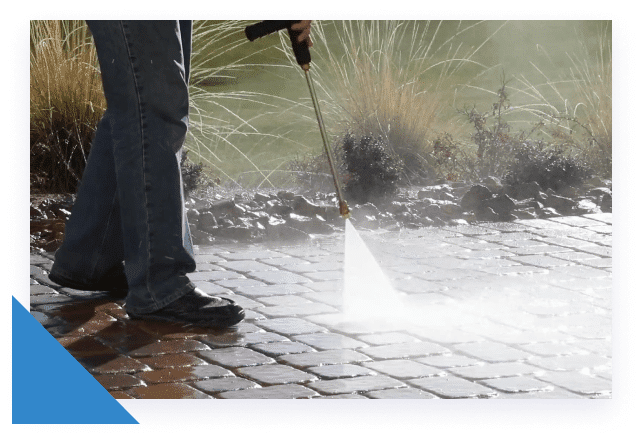 A person in jeans and black shoes uses a pressure washer to clean a brick patio, creating mist.