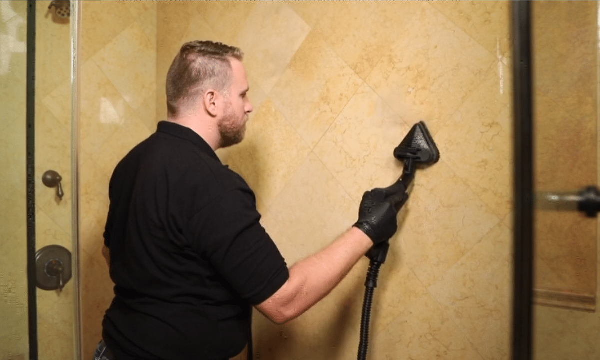 A man in a black shirt and gloves uses a steam cleaner on a beige tiled shower wall.