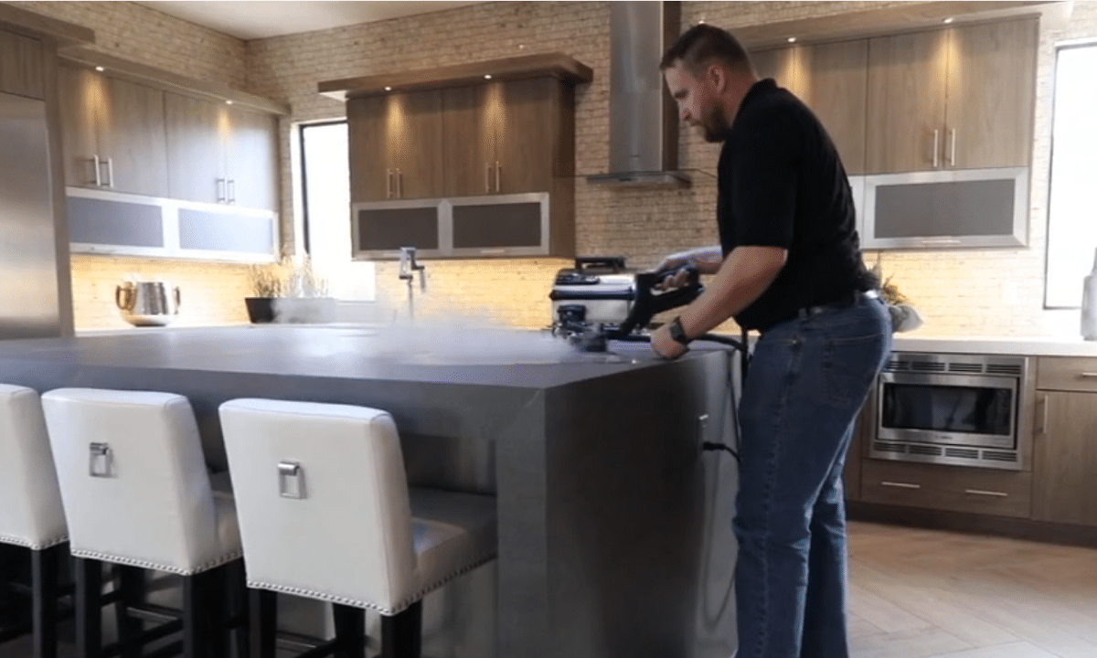 A man in a black shirt and jeans uses a steam cleaner on a kitchen island, producing a cloud of white vapor.