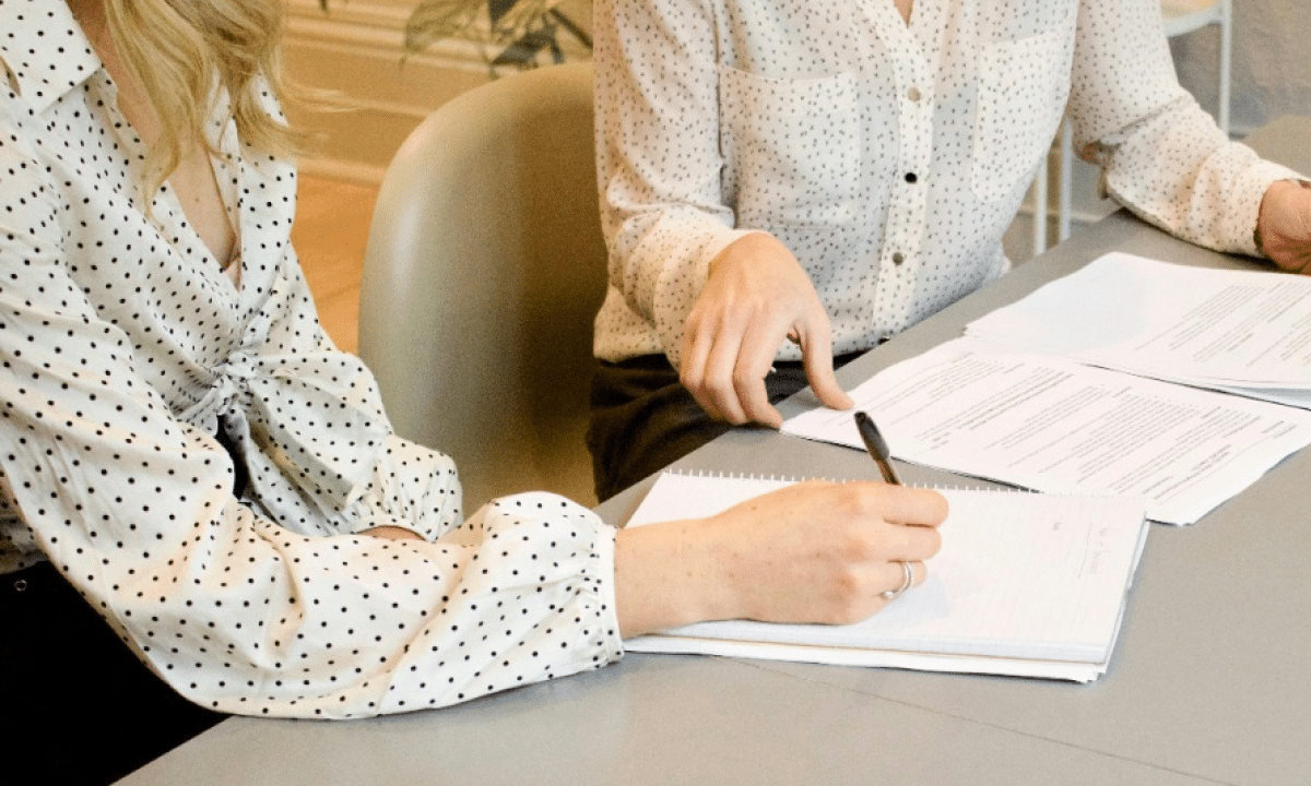 Two women in polka dot shirts sit at a table, one writing in a notebook, the other holding papers.