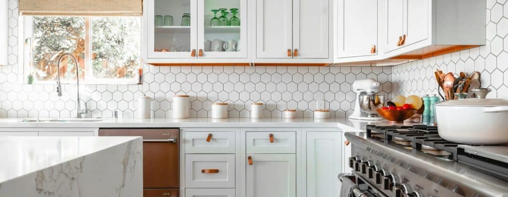 A modern kitchen features white cabinets with leather pulls, a white hexagon tile backsplash, and stainless steel appliances.