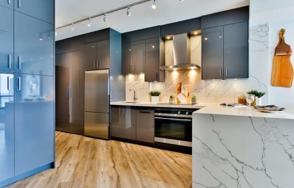 A modern kitchen with light gray cabinets, stainless steel appliances, marble countertops, and light wood flooring.