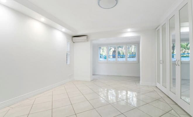 A bright, empty white room with white tiled floors, a window with blinds, and mirrored closet doors.