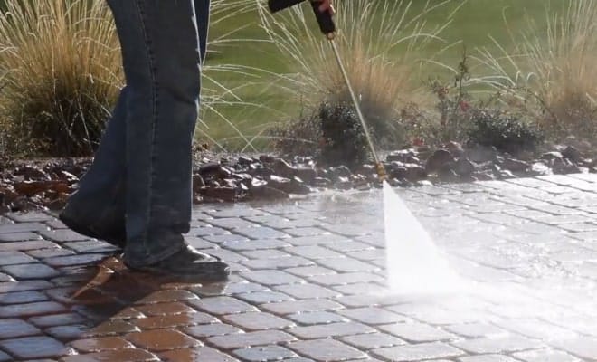 A person in blue jeans and black shoes pressure washes a dirty, brown brick patio, leaving clean, wet bricks.
