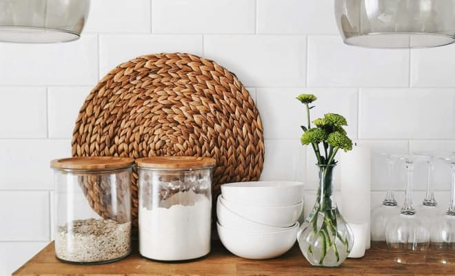 A kitchen counter with two glass jars of dry goods, a woven placemat, white bowls, and green flowers.