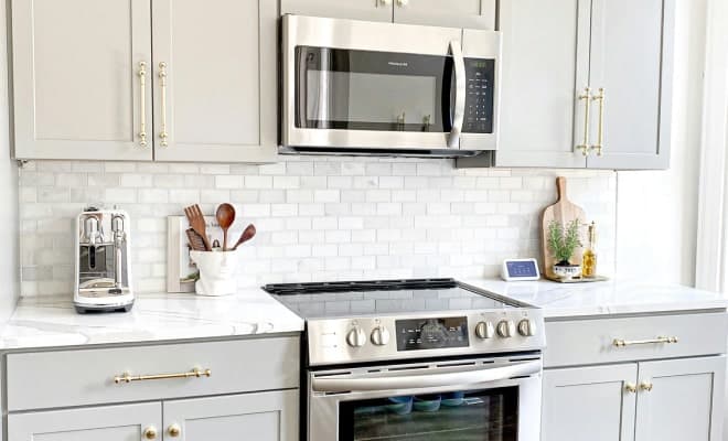 A modern kitchen features light gray cabinets with gold handles, a white subway tile backsplash, and stainless steel appliances, including an "Frigidaire" microwave and an electric range.