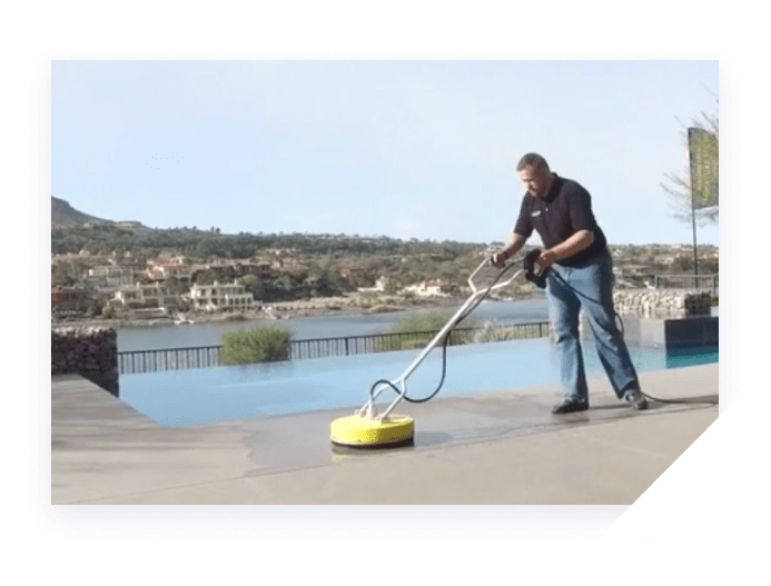 A man in a polo shirt and jeans uses a yellow surface cleaner on a concrete patio next to an infinity pool.
