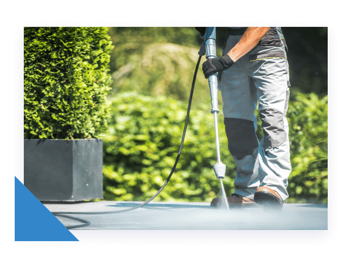 A person in grey work pants and black gloves uses a pressure washer to clean a grey patio.
