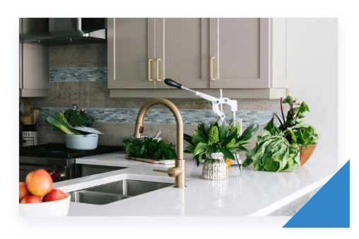 A modern kitchen features light gray cabinets, a white countertop, a brass faucet, and fresh green vegetables.