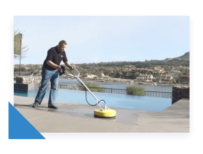 A man in a black shirt and jeans pressure washes a concrete patio next to an infinity pool.
