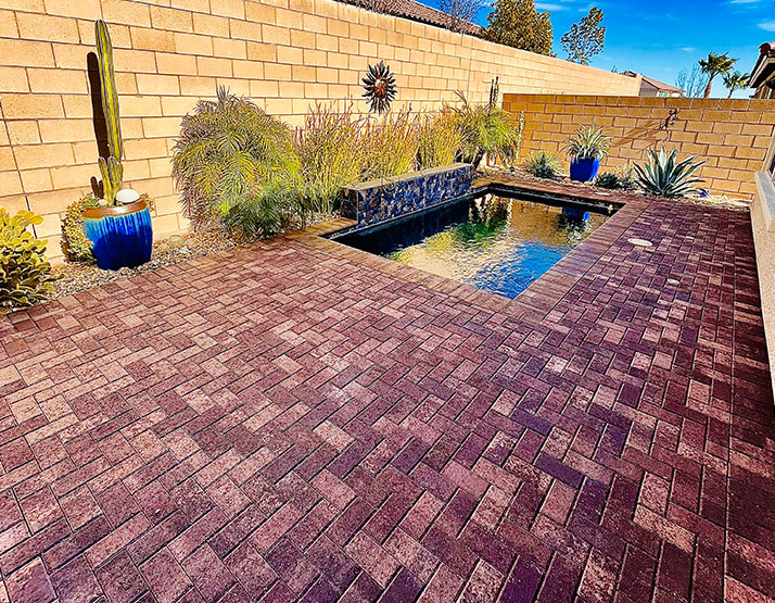 A backyard patio with red brick pavers, a small dark pool, and desert plants against a tan brick wall.