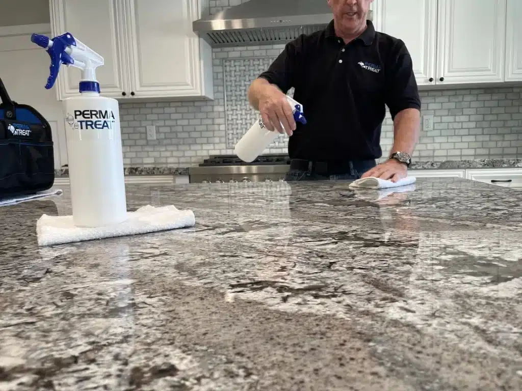 A man in a black shirt sprays and wipes a gray granite countertop in a kitchen with white cabinets.