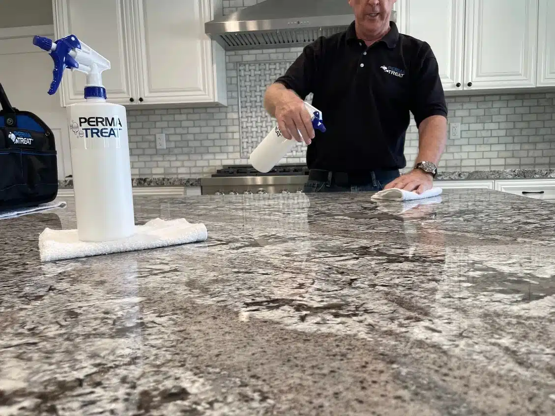A man in a black shirt sprays and wipes a gray granite countertop in a kitchen with white cabinets.