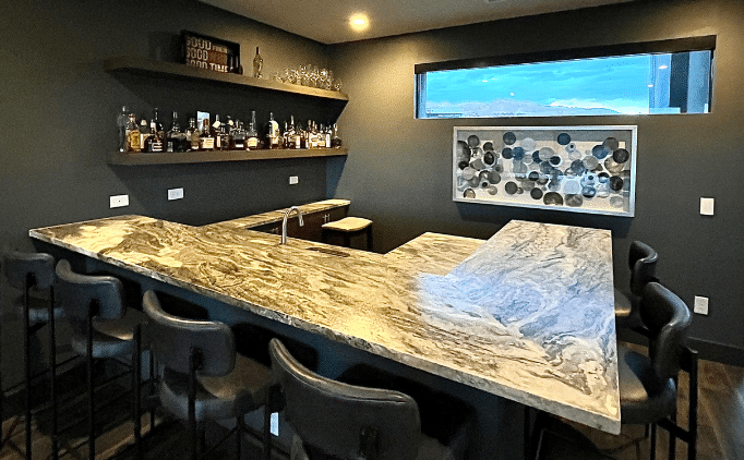 A modern home bar with a dark gray wall, granite countertops, bar stools, and liquor bottles on shelves.