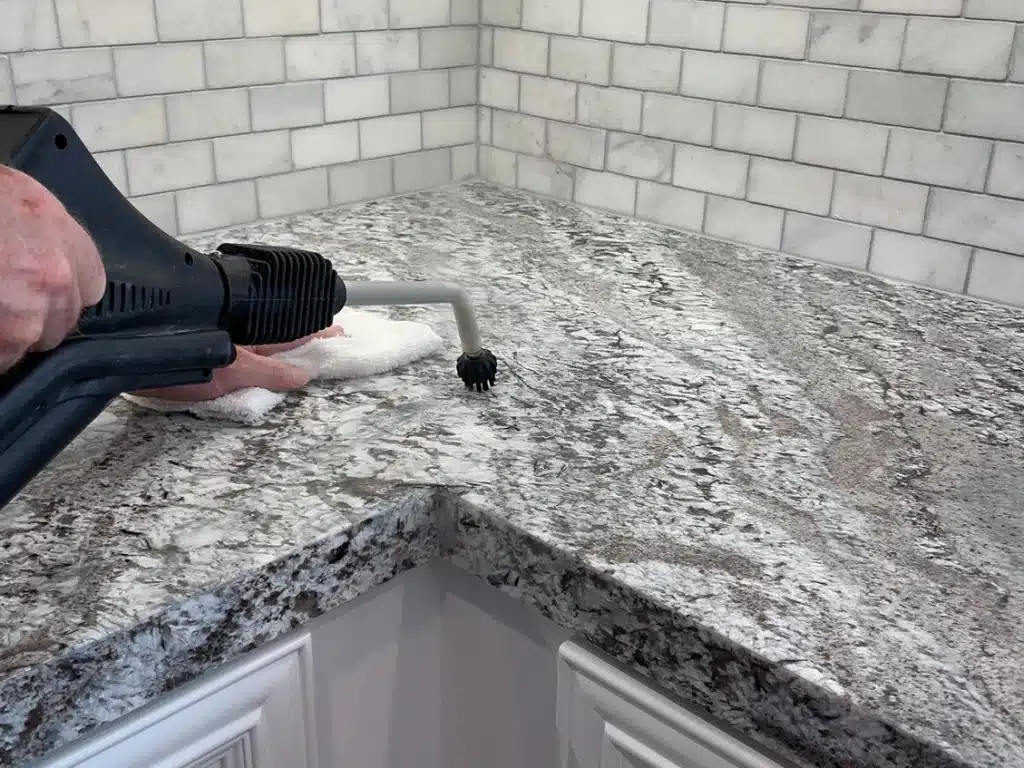 A hand uses a steam cleaner with a small brush attachment on a granite countertop in a kitchen.