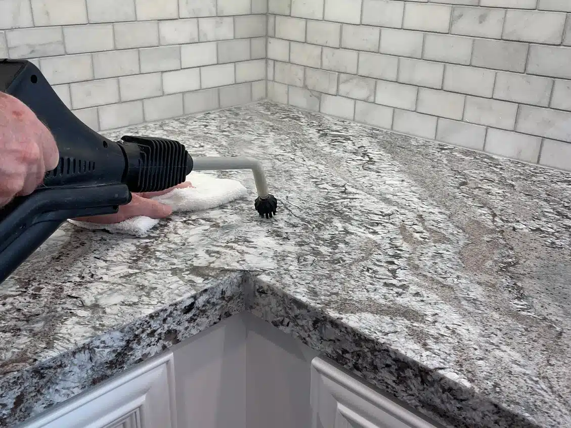 A hand uses a steam cleaner with a small brush attachment on a granite countertop in a kitchen.