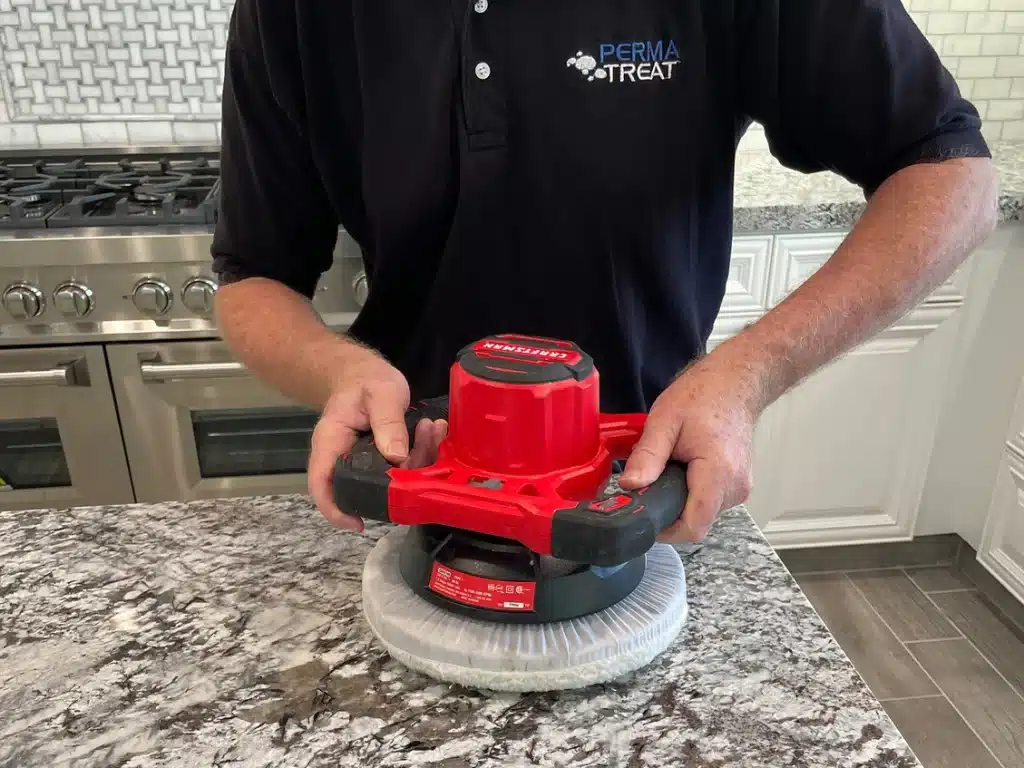 A person in a black "PERMA TREAT" shirt holds a red polisher on a granite countertop in a kitchen.
