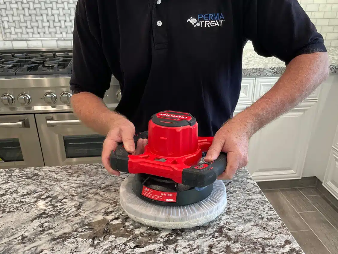 A person in a black "PERMA TREAT" shirt holds a red polisher on a granite countertop in a kitchen.