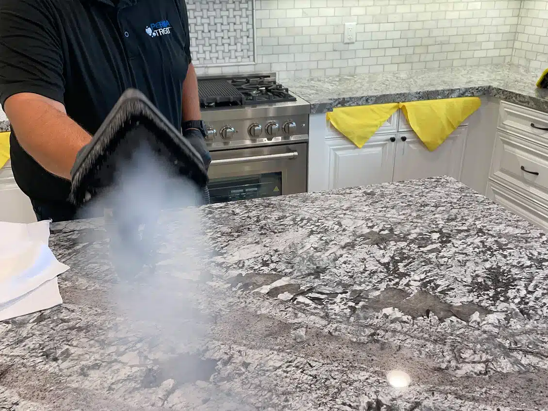 A person in a black shirt with "PERMA TREAT" on it uses a steam cleaner on a granite countertop.