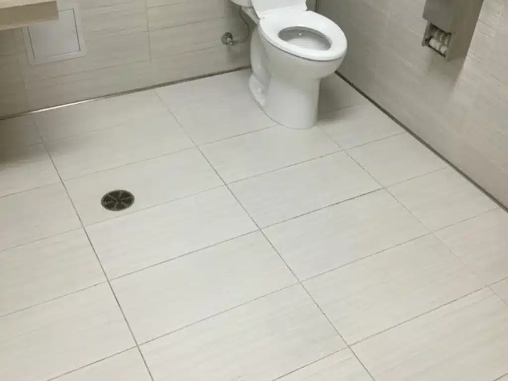 A clean, tiled public restroom with a white toilet, a floor drain, and a toilet paper dispenser.