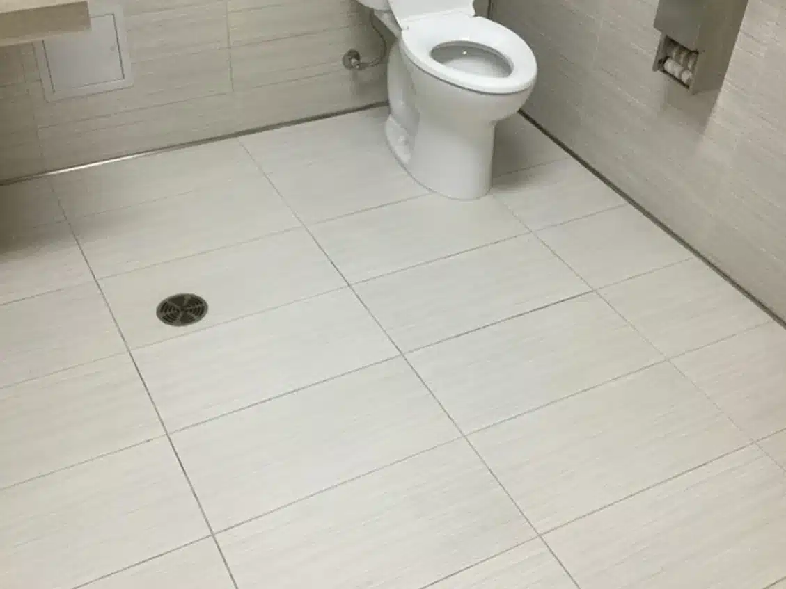 A clean, tiled public restroom with a white toilet, a floor drain, and a toilet paper dispenser.