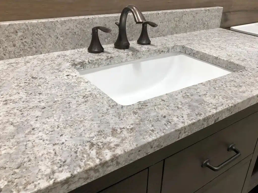 A white rectangular sink is set into a light gray granite countertop with a bronze faucet and handles.