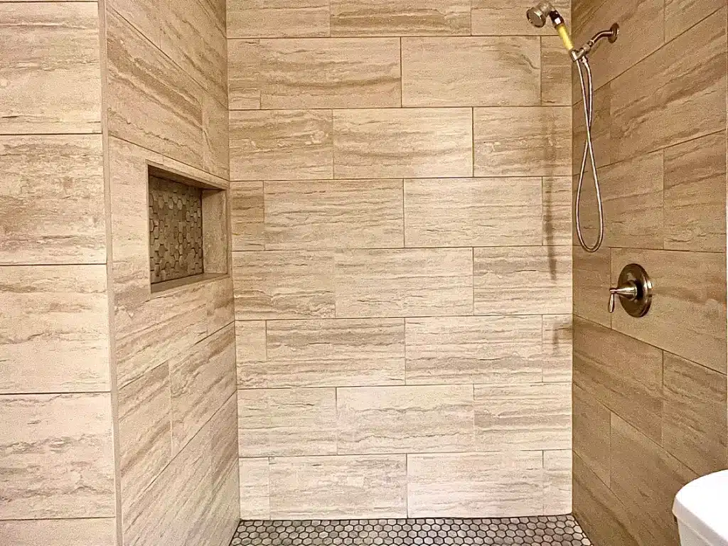 A tiled shower features light brown rectangular tiles, a recessed shelf, a shower head, and a faucet.