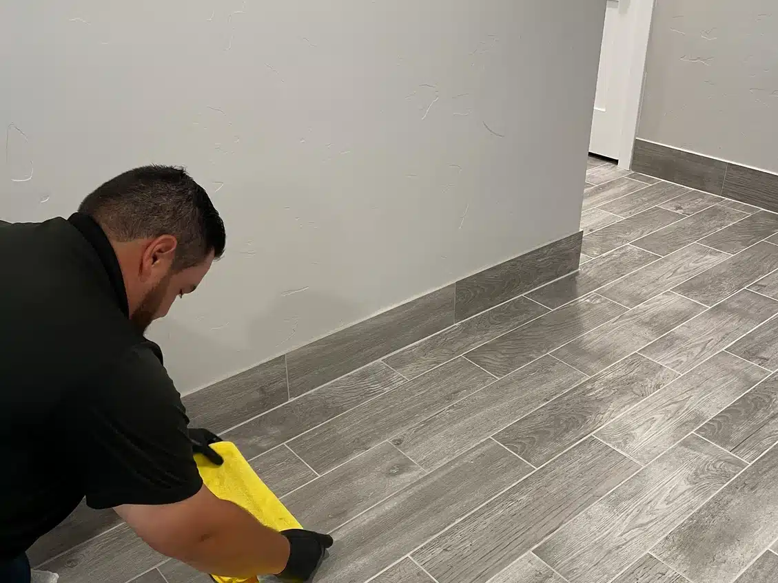 A man in a black shirt and gloves kneels, wiping a gray tiled floor with a yellow cloth.