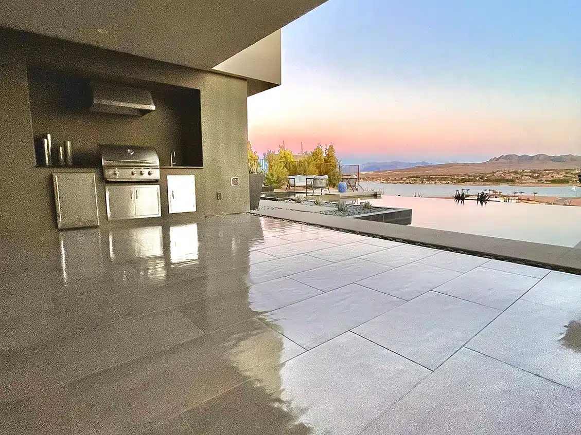 A modern outdoor kitchen with a grill and stainless steel cabinets on a patio overlooking an infinity pool and lake at sunset.