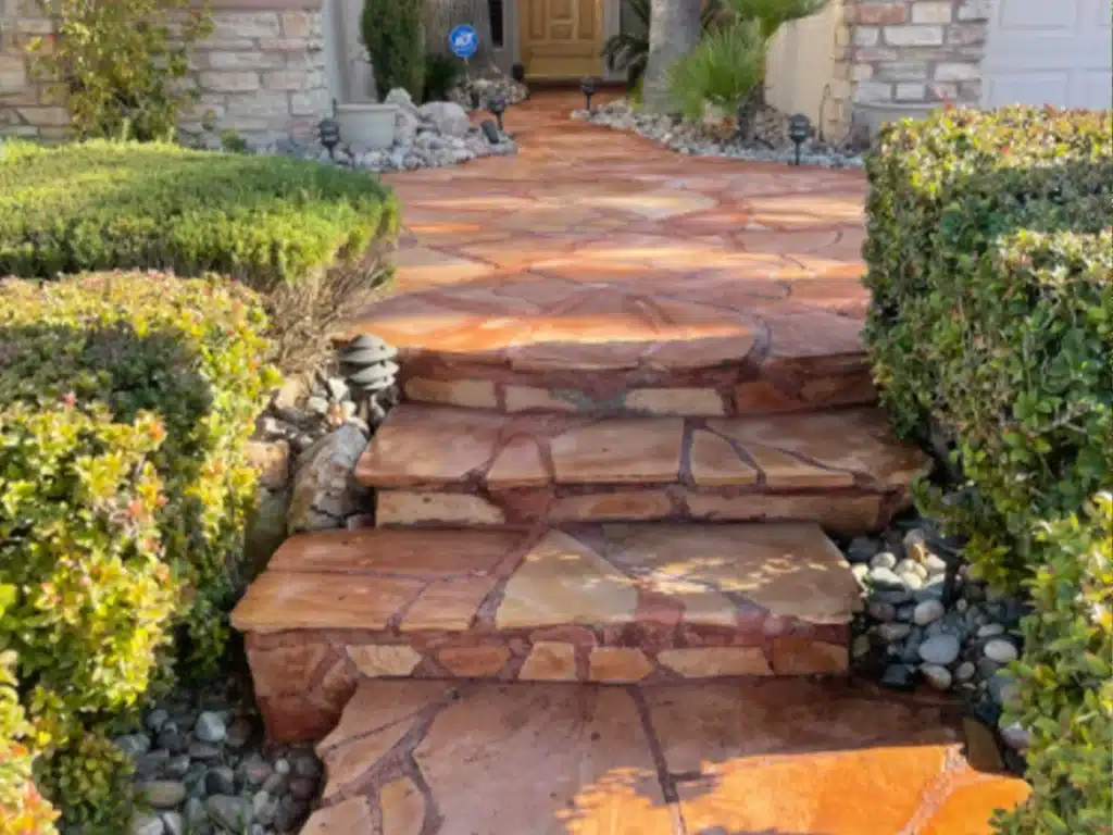 A stone walkway with three steps leads to a house, flanked by green hedges and small gray rocks.